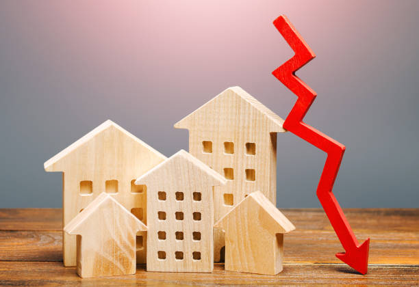 Красная стрелка, опускающаяся над деревянными моделями домов, символизирует снижение рынка недвижимости.