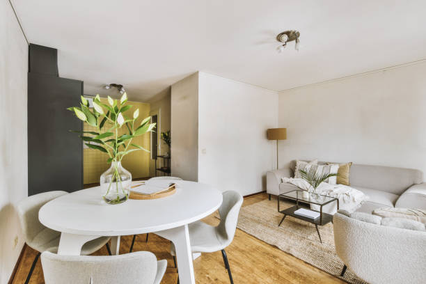 Современная гостиная с обеденной зоной, белым круглым столом с растительным декором, бежевым диваном и минималистским декором.
